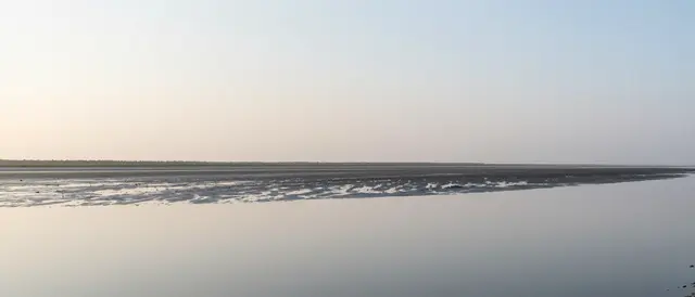 A calm tidal flat reflects the pale soft gradient of the early morning sky beside a distant narrow land strip
