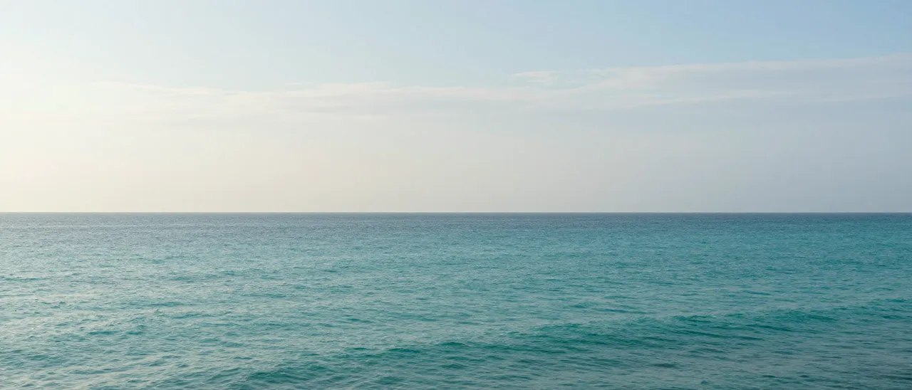 An ultrawide view of a calm turquoise sea meeting a pale sky with thin wispy clouds at the horizon