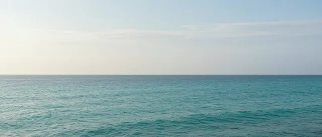 An ultrawide view of a calm turquoise sea meeting a pale sky with thin wispy clouds at the horizon