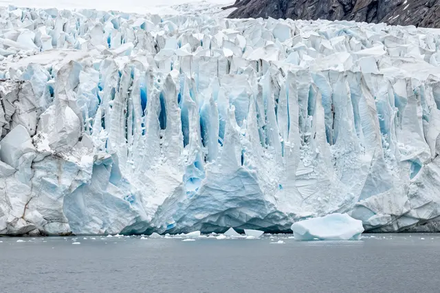 Towering jagged glacier front meets calm gray fjord water with floating ice chunks