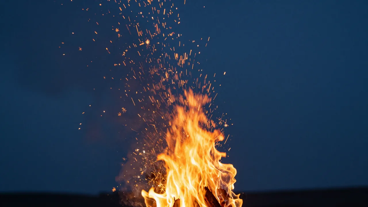 Bright campfire flames send glowing sparks rising upward against a dark deep blue night sky