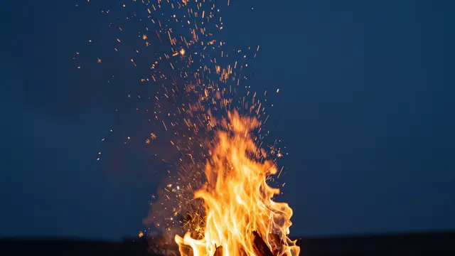 Bright campfire flames send glowing sparks rising upward against a dark deep blue night sky