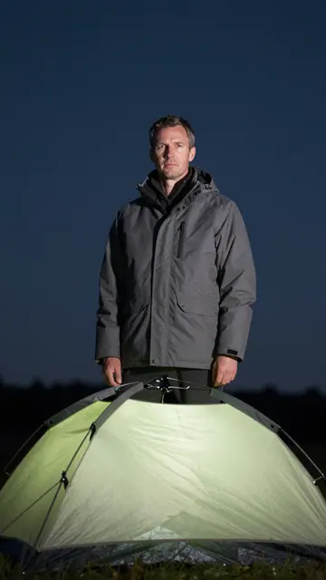 A man in a gray parka stands behind a glowing dome tent under a dark night sky