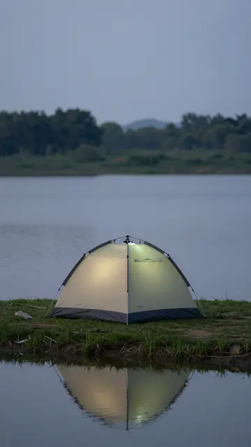 Lit dome tent on a small grassy lakeside patch with still water reflections at twilight