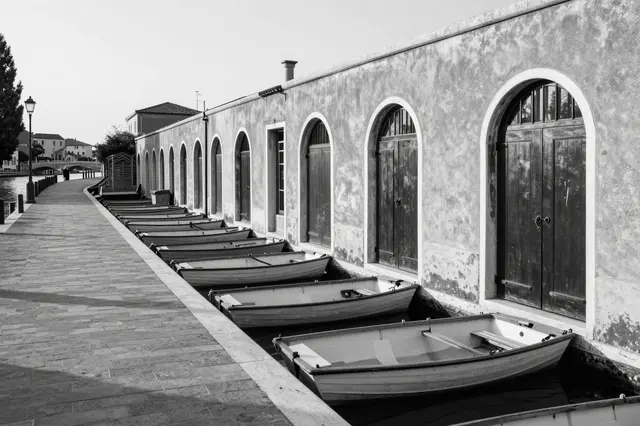 Black and white dawn view of a stone canal walkway lined with moored rowboats beside arched doorways