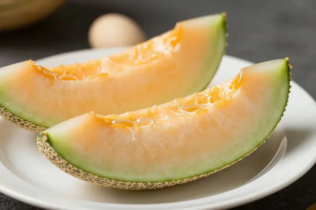 Two ripe cantaloupe wedges rest on a plain white ceramic plate against a dark neutral background