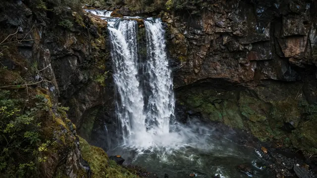 A waterfall cascades over a rugged mossy rock cliff into a deep turbulent canyon pool