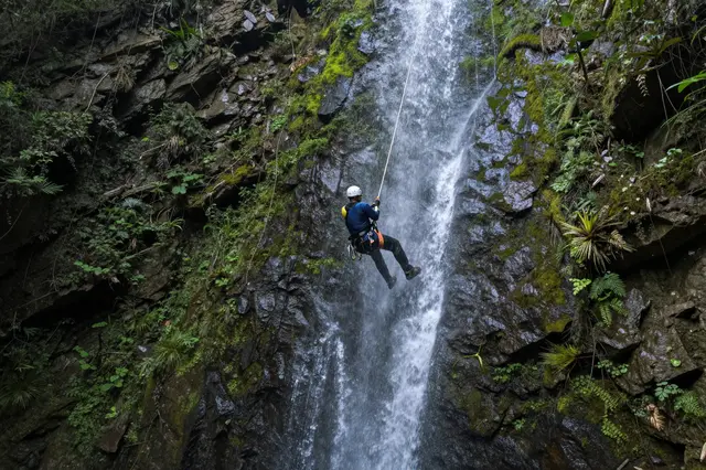A canyoneer in a white helmet rappels on a rope beside a tall rocky waterfall covered in green moss