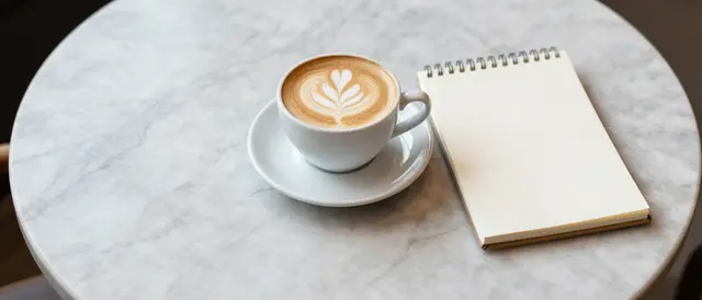Top down view of a latte art cappuccino and blank spiral notepad resting on a round marble cafe table with empty copy space