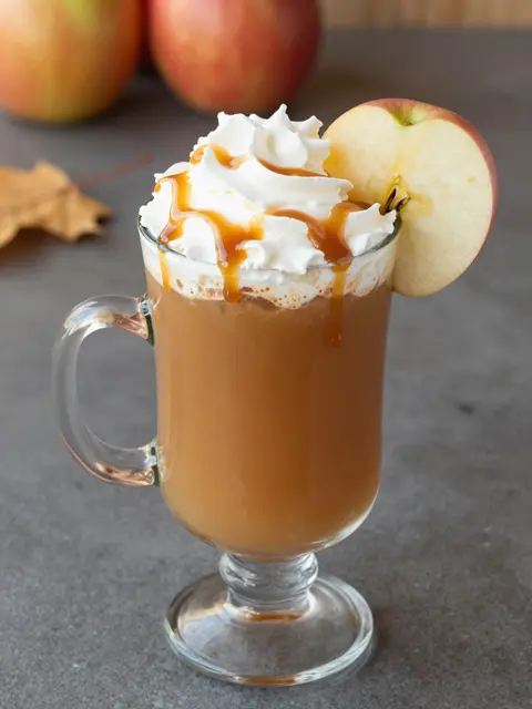 Tall glass mug of caramel apple cider topped with whipped cream, caramel drizzle and an apple slice on the rim, with autumn