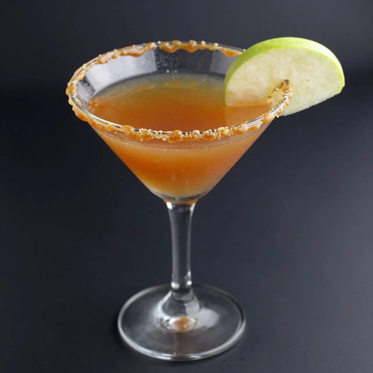 Caramel apple martini in a martini glass with a caramel drizzled rim and green apple garnish against a dark background