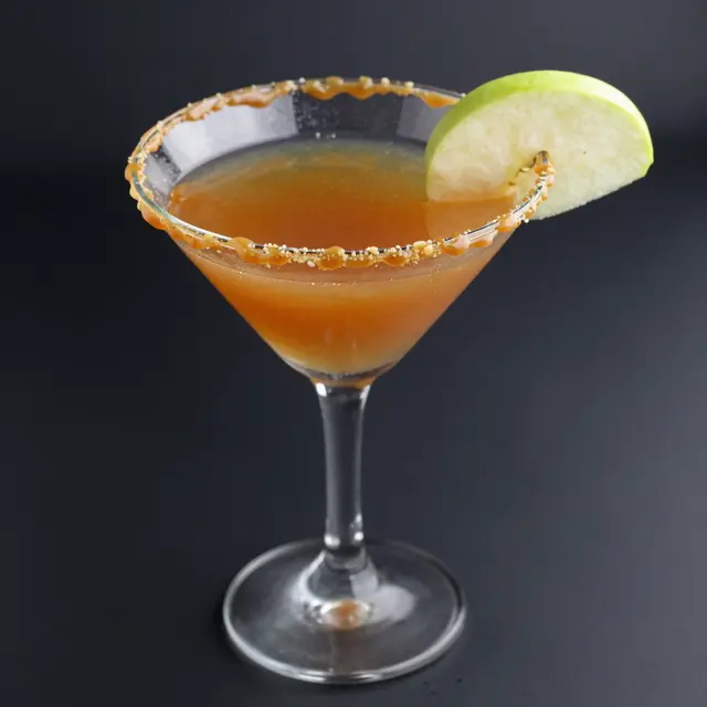 Caramel apple martini in a martini glass with a caramel drizzled rim and green apple garnish against a dark background