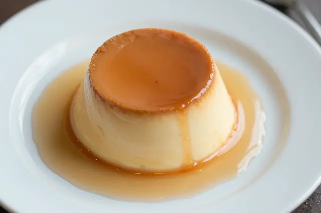 A smooth caramel flan sits on a white plate with golden syrup pooling around its base.
