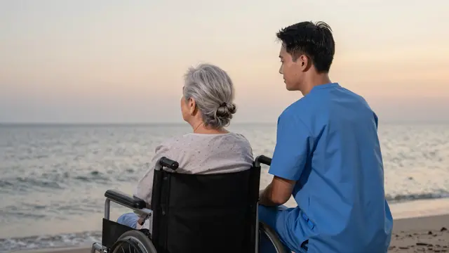 A male caregiver and an older woman in a wheelchair sit on a beach watching the calm sea at sunset, viewed from behind.