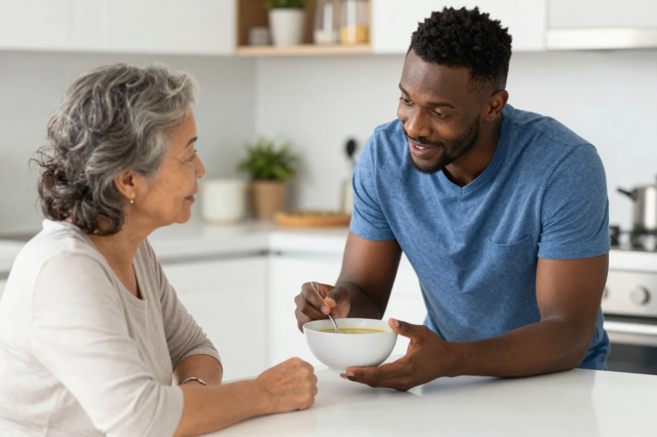 A male caregiver offers a bowl of soup to an older East Asian woman at a bright kitchen table