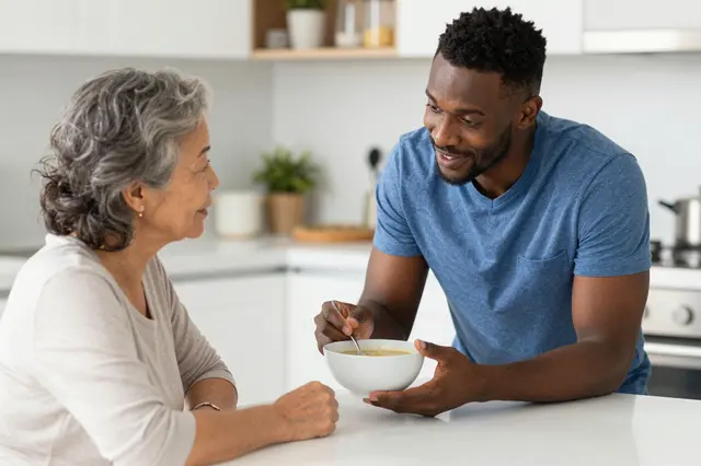 A male caregiver offers a bowl of soup to an older East Asian woman at a bright kitchen table