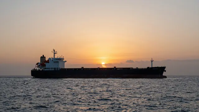 A bulk cargo vessel floats on calm open sea beneath a soft orange setting sun