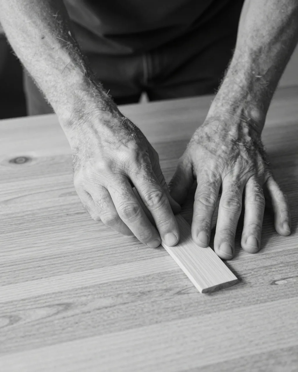 Monochrome close-up of an older carpenter's hands inspecting a small wood piece on a tabletop