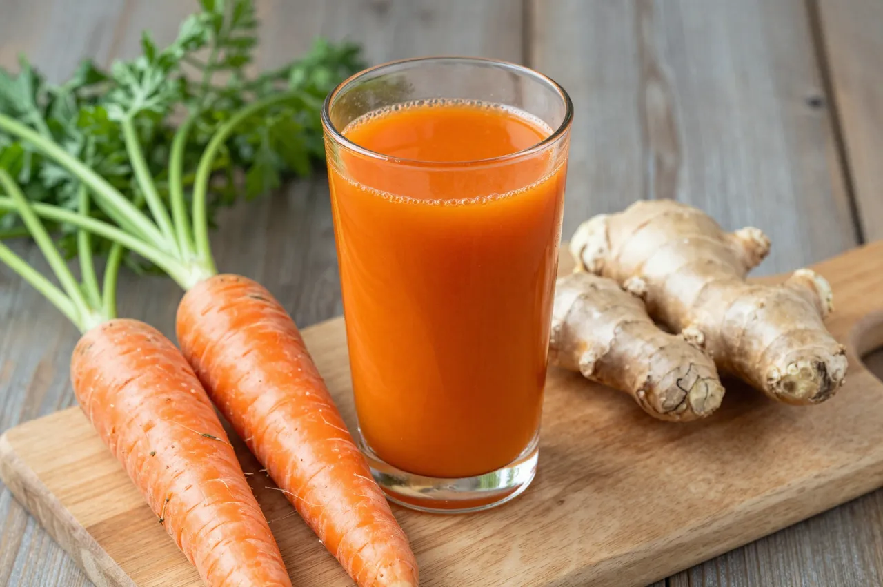 A glass of bright orange carrot juice sits on a wooden board with whole fresh carrots and ginger root.