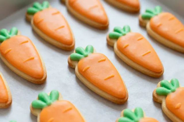 Rows of decorated carrot shaped sugar cookies with orange icing and green tops rest on baking parchment.
