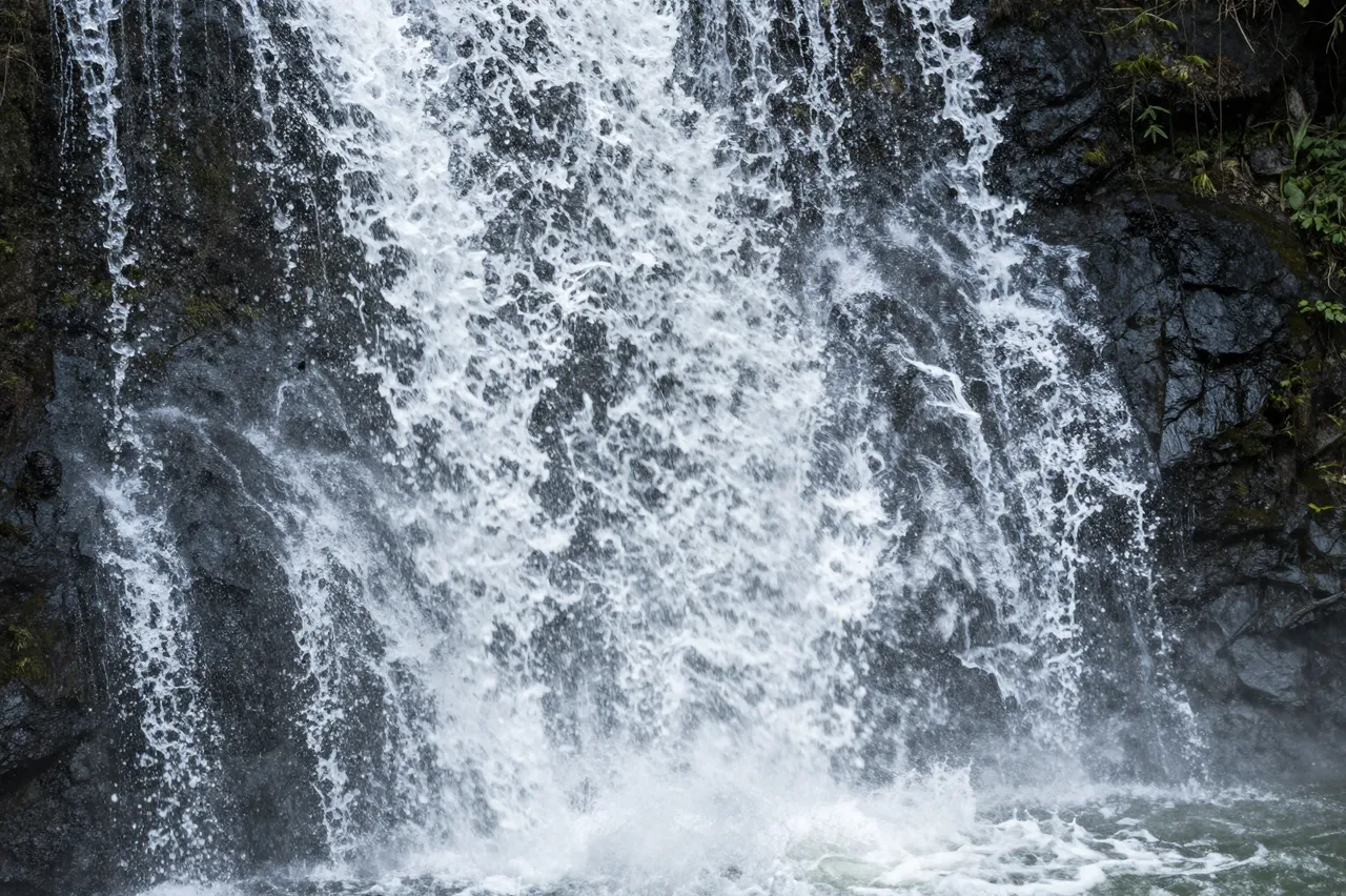 Turbulent white waterfall pours over dark wet mossy rock into a churning pool below