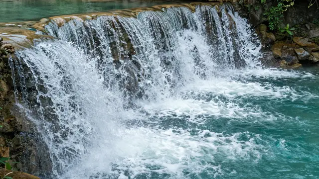 A tiered cascading waterfall flows over rocky ledges into a bright turquoise natural pool