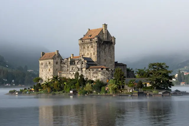 A historic stone castle sits on a small tree-covered island surrounded by calm misty lake water