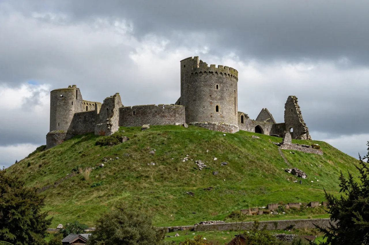 Medieval stone castle ruins sit atop a lush green hill under dramatic storm clouds