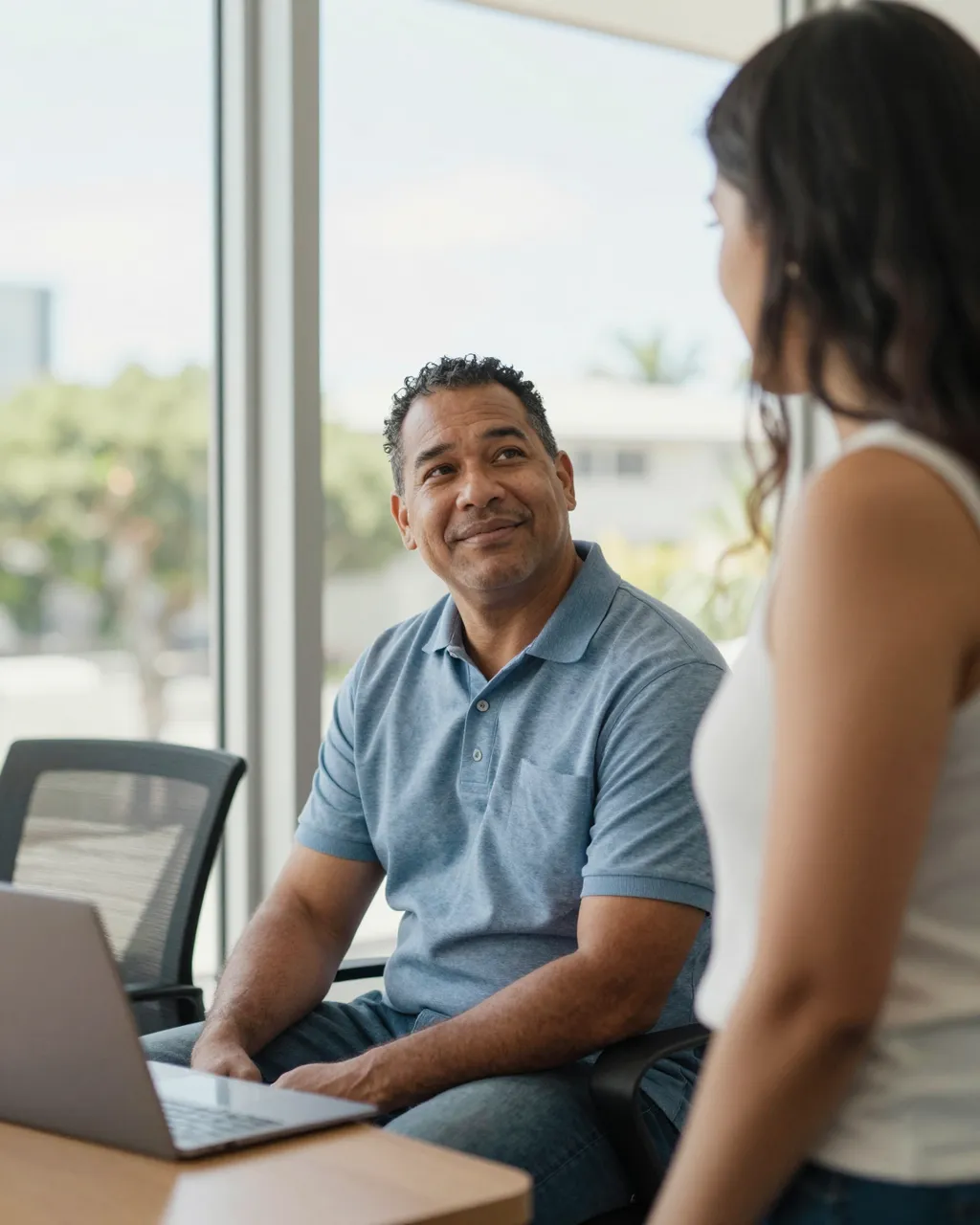 Two coworkers converse beside a sunny office window next to a laptop on a desk