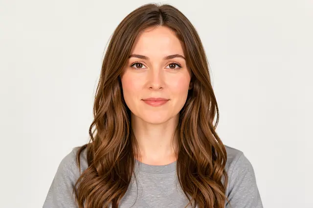 A woman with long wavy brown hair wears a gray shirt and looks directly at the camera against a plain light background.