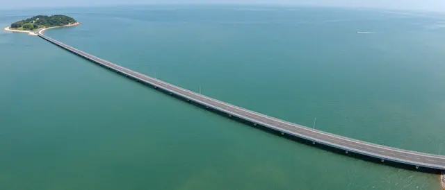 Aerial view of a long paved causeway crossing turquoise sea to a small forested island