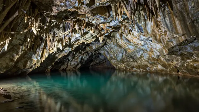A rocky cave with hanging stalactites holds a calm turquoise underground lake that reflects the cavern walls