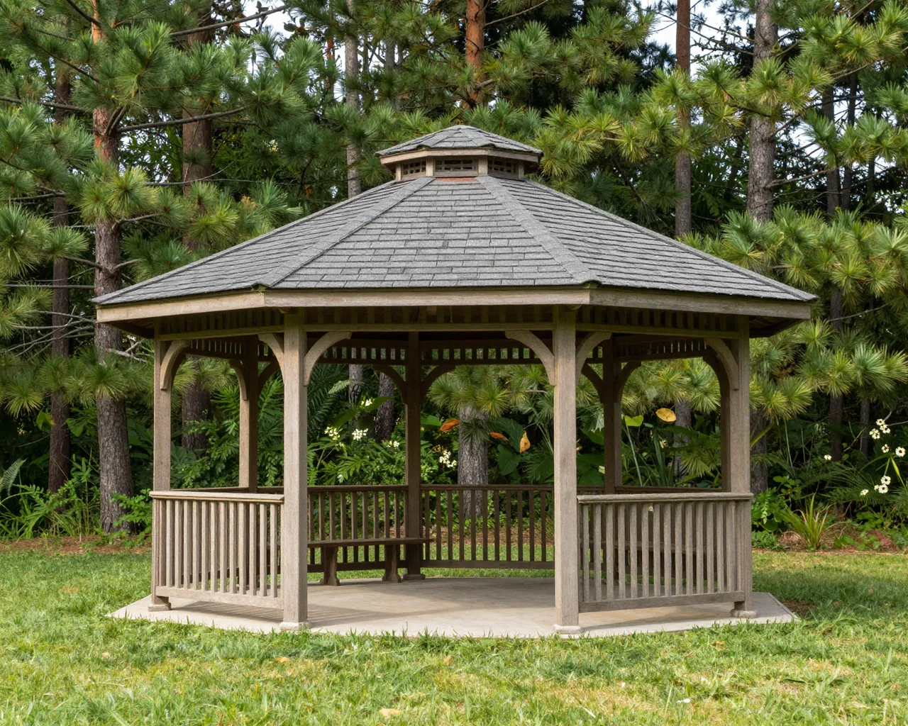 A wooden cedar gazebo with a shingled roof sits on a lawn surrounded by tall pine trees