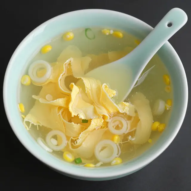 Overhead view of egg drop soup with corn, scallions and a ceramic spoon in a pale green bowl on a dark surface