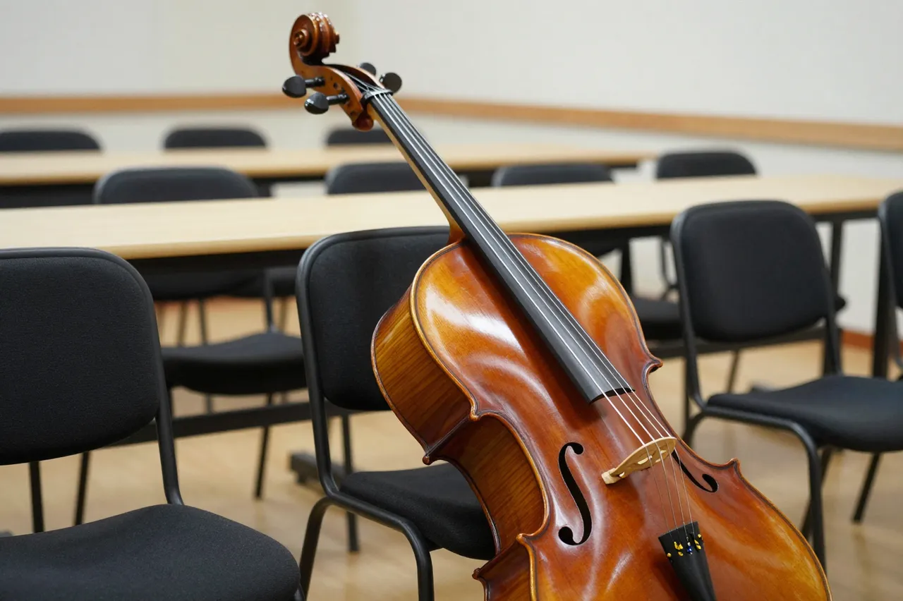 A polished wooden cello leans against an empty black upholstered chair in a sparse rehearsal room.