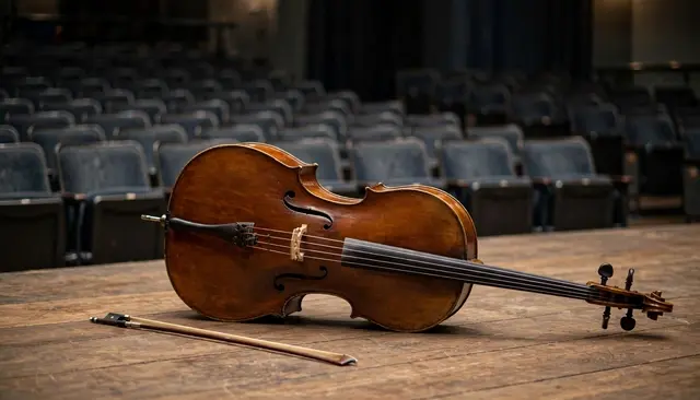 A wooden cello and bow rest on their side on a worn stage floor, with empty theater seats blurred in the background.
