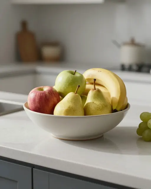 A white ceramic fruit bowl filled with apples, pears, and bananas sits on a bright kitchen countertop beside green grapes.