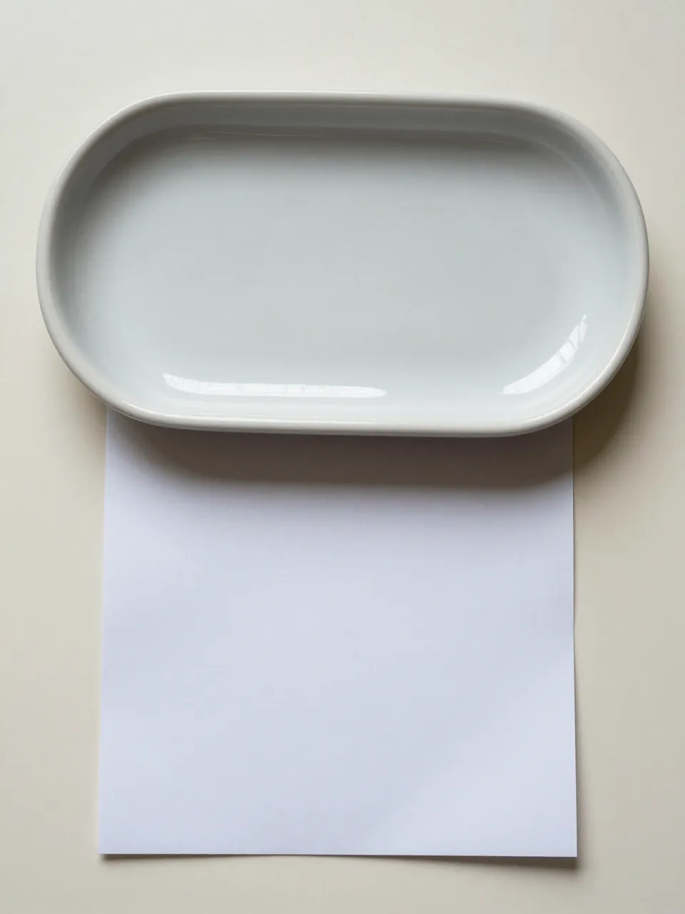 Top down view of a light gray ceramic oval tray resting above a blank white sheet of paper on a cream surface with copy