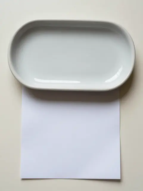Top down view of a light gray ceramic oval tray resting above a blank white sheet of paper on a cream surface with copy