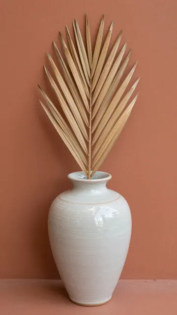 A ribbed white ceramic vase holding a dried palm leaf stands against a smooth terracotta wall.