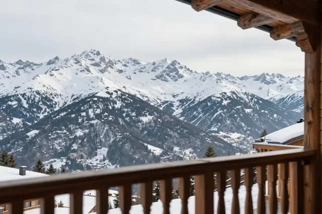 View from a wooden chalet balcony overlooking a range of snow covered alpine peaks under an overcast sky.