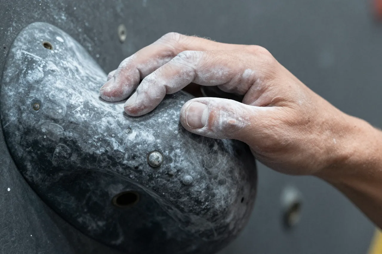 Close-up of a chalk-covered hand gripping a dark climbing hold on an indoor climbing wall