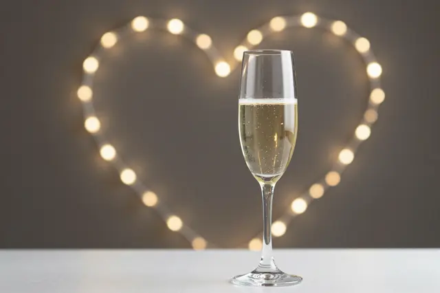A filled champagne flute sits on a light table in front of a heart-shaped string light bokeh background