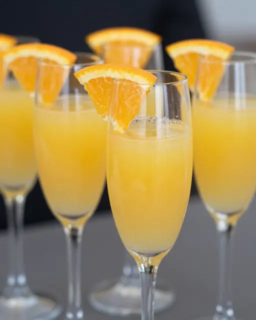A close-up of multiple filled champagne flutes garnished with fresh orange slices arranged on a table.