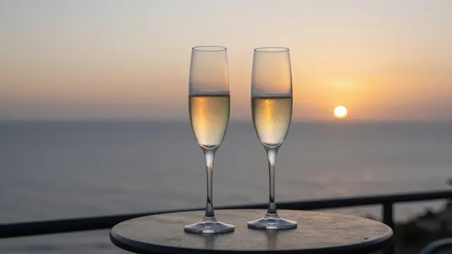 Two filled champagne flutes rest on a small balcony table overlooking a calm sea at golden sunset