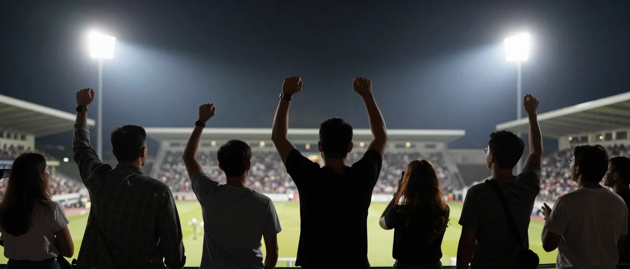 Group of cheering sports fans stand in stadium stands under bright floodlights watching a night match