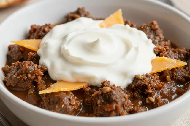 A close-up view of a bowl of chili con carne topped with swirled sour cream and cheddar cheese shards