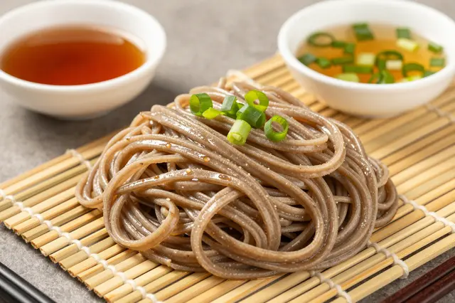 Chilled buckwheat soba noodles rest on a bamboo mat alongside small bowls of dipping broth and sliced scallions