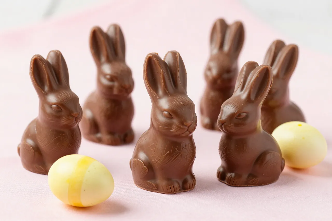Unwrapped milk chocolate bunnies and candy eggs are arranged on a soft pastel pink fabric surface for Easter.