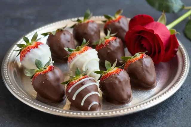 Chocolate dipped strawberries arranged on a silver tray beside a single red rose on a dark textured background